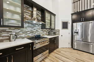 Kitchen with high end appliances, wall chimney exhaust hood, decorative backsplash, light wood-type flooring, and glass insert cabinets