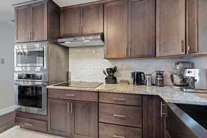 Kitchen featuring stainless steel appliances, ventilation hood, light stone counters, dark brown cabinetry, and decorative backsplash