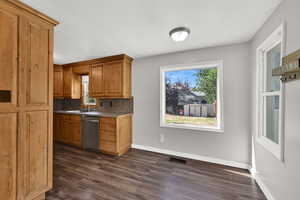 Kitchen featuring dark wood-style floors, tasteful backsplash, stainless steel dishwasher, brown cabinets, and dark countertops