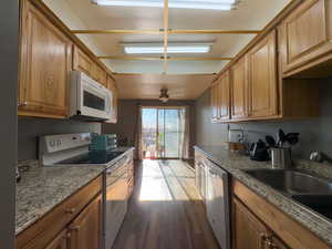 Kitchen featuring white appliances, wood finished floors, light stone countertops, brown cabinetry, and a ceiling fan