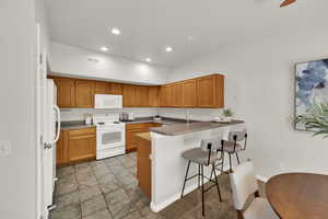 Kitchen featuring recessed lighting, white appliances, a peninsula, a kitchen bar, and brown cabinetry