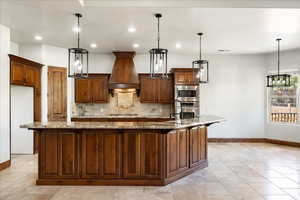 Kitchen featuring light stone countertops, a large island with sink, custom exhaust hood, backsplash, and appliances with stainless steel finishes