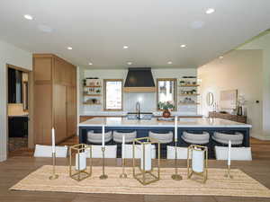 Kitchen with open shelves, dark wood-style flooring, a center island with sink, premium range hood, and recessed lighting