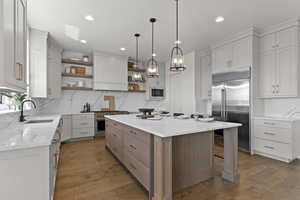 Kitchen with open shelves, wood finished floors, a center island, light stone countertops, and built in appliances