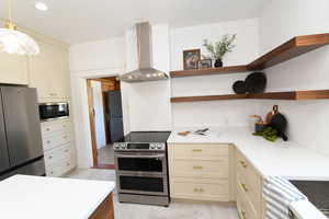 Kitchen featuring open shelves, appliances with stainless steel finishes, wall chimney range hood, cream cabinetry, and light marble finish flooring
