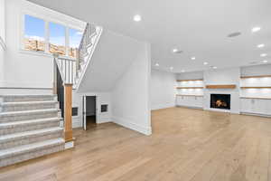 Unfurnished living room featuring light wood-style flooring, stairway, recessed lighting, a warm lit fireplace, and built in shelves