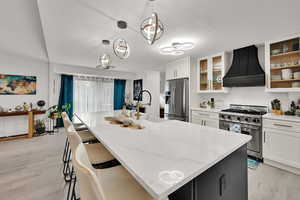 Kitchen with premium range hood, appliances with stainless steel finishes, light wood-style floors, light stone counters, and white cabinets