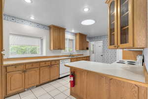 Kitchen with wallpapered walls, glass insert cabinets, a peninsula, white appliances, and light countertops