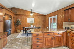 Kitchen featuring wooden walls, vaulted ceiling, a peninsula, ceiling fan, and brown cabinetry