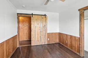 Unfurnished bedroom with a barn door, hardwood / wood-style floors, wooden walls, a wainscoted wall, and ornamental molding