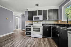 Kitchen with dark cabinetry, ornamental molding, white appliances, dark countertops, and wood finished floors