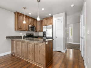 Kitchen with a peninsula, dark wood-style floors, dark stone counters, brown cabinetry, and appliances with stainless steel finishes