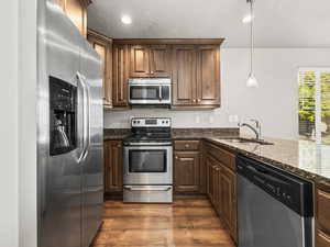 Kitchen featuring appliances with stainless steel finishes, dark wood-type flooring, dark stone counters, decorative light fixtures, and a textured ceiling