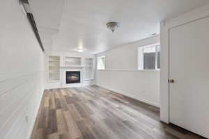 Unfurnished living room featuring built in shelves, wood finished floors, a glass covered fireplace, and a wainscoted wall