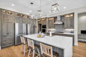 Kitchen featuring light wood-style flooring, wall chimney exhaust hood, tasteful backsplash, stainless steel appliances, and a kitchen breakfast bar
