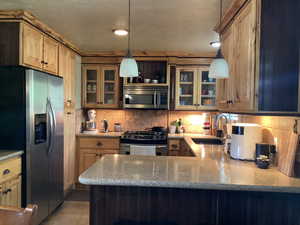 Kitchen with appliances with stainless steel finishes, tasteful backsplash, a peninsula, hanging light fixtures, and glass insert cabinets