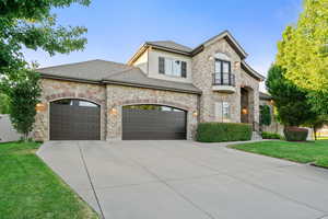 French country style house with a balcony, stone siding, a front lawn, driveway, and roof with shingles