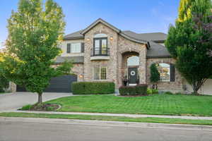French country inspired facade with a balcony, concrete driveway, an attached garage, stone siding, and a front lawn
