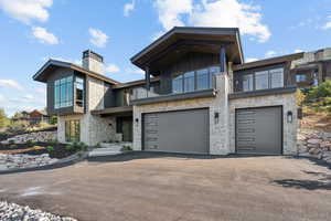Modern home featuring a balcony, asphalt driveway, stone siding, a chimney, and a garage