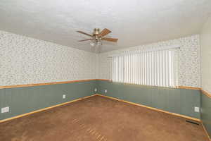 Carpeted empty room with wallpapered walls, a textured ceiling, ceiling fan, and a wainscoted wall