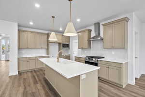 Kitchen featuring wall chimney exhaust hood, gas range, an island with sink, tasteful backsplash, and pendant lighting