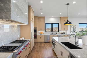 Kitchen featuring light brown cabinets, light stone counters, wall chimney exhaust hood, appliances with stainless steel finishes, and backsplash