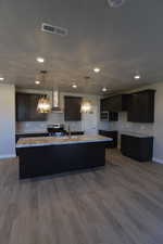 Kitchen featuring a center island with sink, stainless steel range with gas stovetop, wood finished floors, light stone counters, and recessed lighting