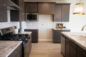 Kitchen featuring appliances with stainless steel finishes, dark brown cabinets, wall chimney exhaust hood, and light stone countertops