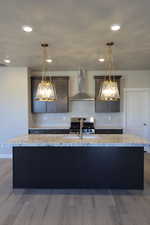Kitchen with stainless steel stove, wall chimney exhaust hood, a kitchen island with sink, recessed lighting, and light stone countertops
