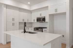 Kitchen with light wood finished floors, white cabinets, stainless steel appliances, light stone countertops, and an island with sink