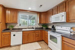 Kitchen featuring white appliances, brown cabinetry, dark countertops, and light tile patterned floors