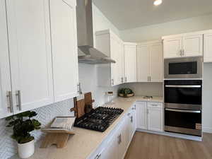 Kitchen with wall chimney exhaust hood, appliances with stainless steel finishes, decorative backsplash, white cabinetry, and light wood-style floors