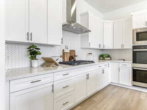 Kitchen featuring appliances with stainless steel finishes, white cabinets, backsplash, wall chimney range hood, and light wood-style flooring