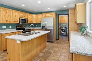 Kitchen with appliances with stainless steel finishes, washing machine and dryer, recessed lighting, a kitchen island with sink, and light stone countertops