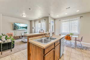 Kitchen with light tile patterned floors, brown cabinets, a center island with sink, stainless steel dishwasher, and light countertops