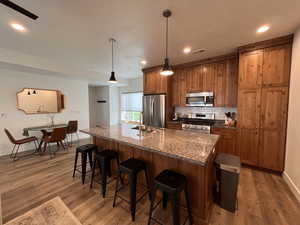 Kitchen with brown cabinetry, appliances with stainless steel finishes, a center island with sink, a kitchen bar, and wood finished floors