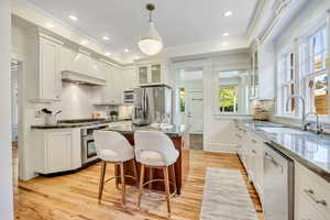 Kitchen featuring backsplash, premium range hood, stainless steel appliances, dark stone counters, and ornamental molding