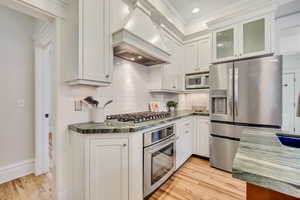 Kitchen featuring stainless steel appliances, decorative backsplash, ornamental molding, premium range hood, and light wood finished floors