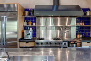 Detailed view of stainless steel countertops, high end appliances, backsplash, open shelves, and exhaust hood