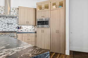 Kitchen featuring light brown cabinetry, wall chimney range hood, decorative backsplash, appliances with stainless steel finishes, and dark wood-type flooring