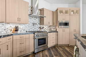 Kitchen featuring light brown cabinetry, wall chimney exhaust hood, appliances with stainless steel finishes, dark wood finished floors, and dark stone counters