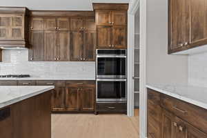 Kitchen with backsplash, double oven, light wood-style flooring, light stone counters, and dark brown cabinets