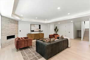 Living room with a stone fireplace, light wood-type flooring, recessed lighting, stairs, and a raised ceiling