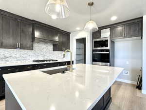 Kitchen featuring light wood finished floors, backsplash, appliances with stainless steel finishes, decorative light fixtures, and light stone countertops
