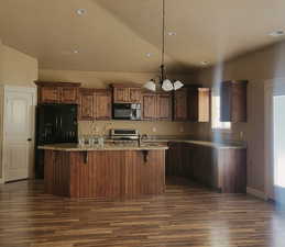 Kitchen featuring granite counters, dark wood cabinets, lofted ceiling, black appliances, a kitchen island with deep double s.s. sink, dark wood-style floors, and pendant lighting