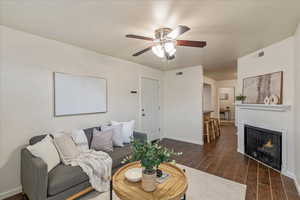 Living area with dark wood-style tile floors, a warm lit fireplace, and ceiling fan