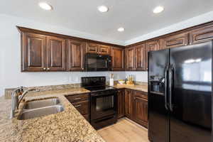Kitchen featuring black appliances, light wood finished floors, recessed lighting, light stone countertops, and dark brown cabinets
