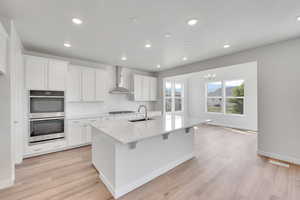 Kitchen featuring a center island with sink, stainless steel appliances, light countertops, wall chimney range hood, and white cabinetry