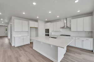 Kitchen featuring decorative backsplash, wall chimney range hood, recessed lighting, white cabinets, and a center island with sink