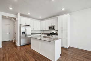 Kitchen featuring appliances with stainless steel finishes, dark wood-style floors, a textured ceiling, a kitchen island with sink, and white cabinetry
